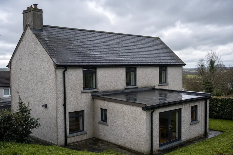 A rendered Irish-style house with a dark slate pitched roof and a small rear extension with a wet flat roof.
