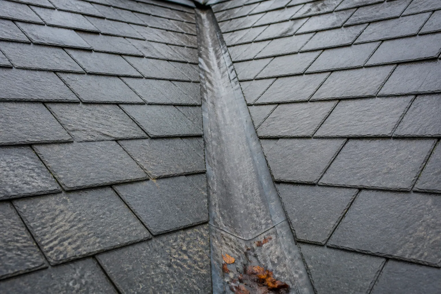 Weathered lead-lined valley between two damp grey slate roof slopes, with a few autumn leaves near the bottom.