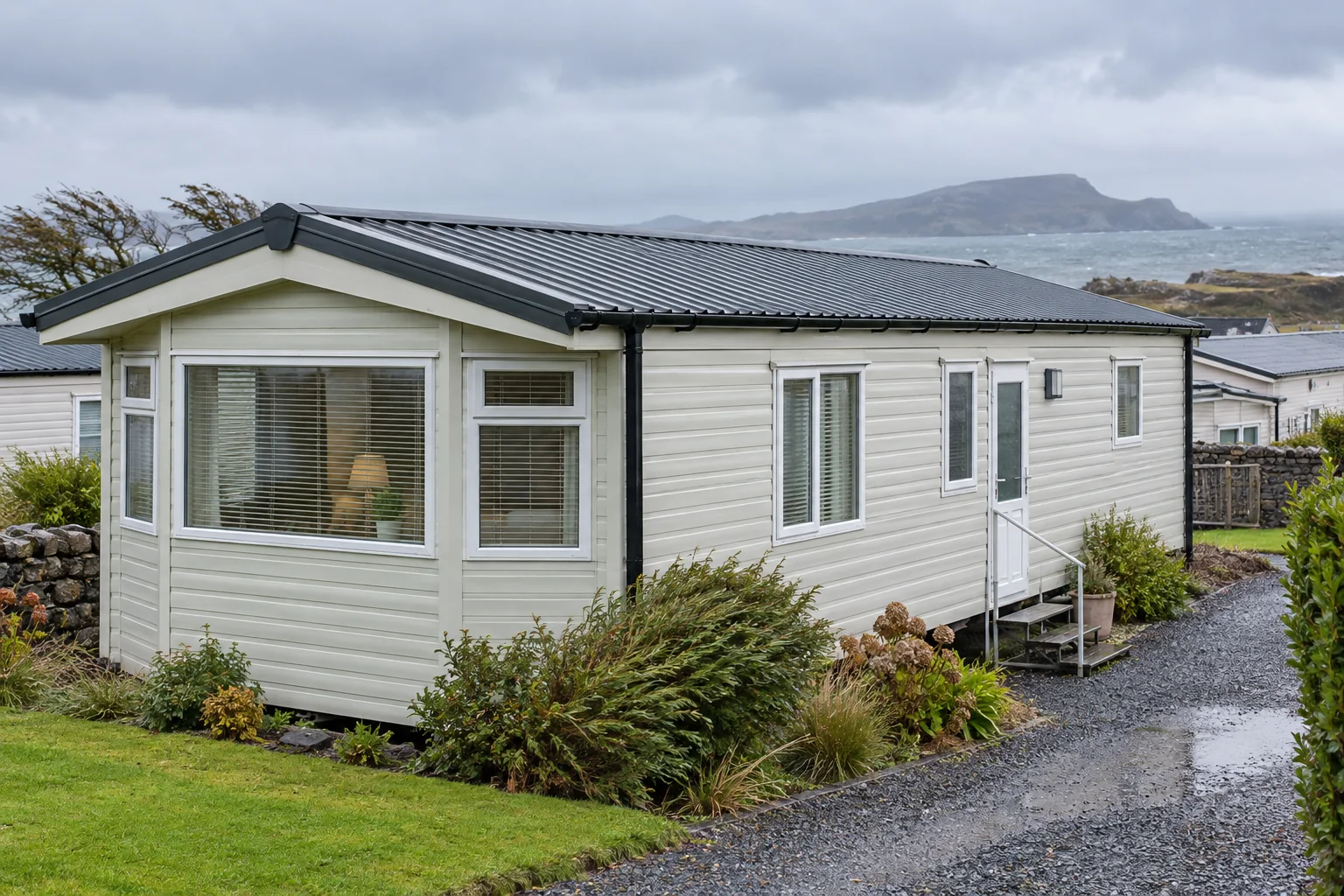 A residential mobile home with a shallow metal-clad roof and black gutters in wet, windy Irish coastal weather.